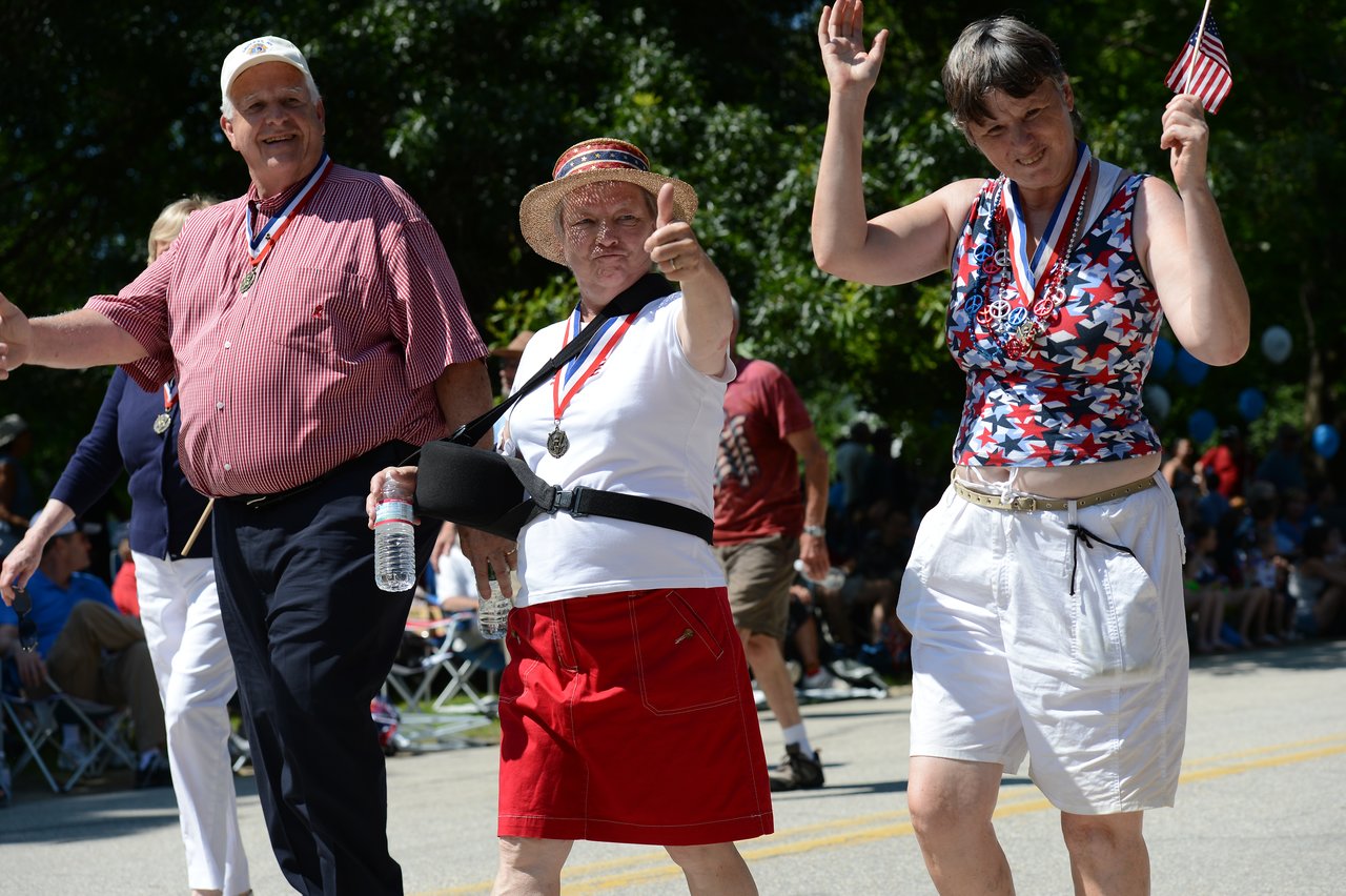 Three people wearing medals walk in a parade, smiling and waving.