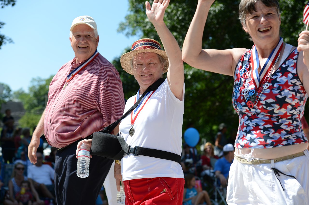 Three people wearing patriotic outfits and medals smile and wave while walking in a Fourth of July parade.