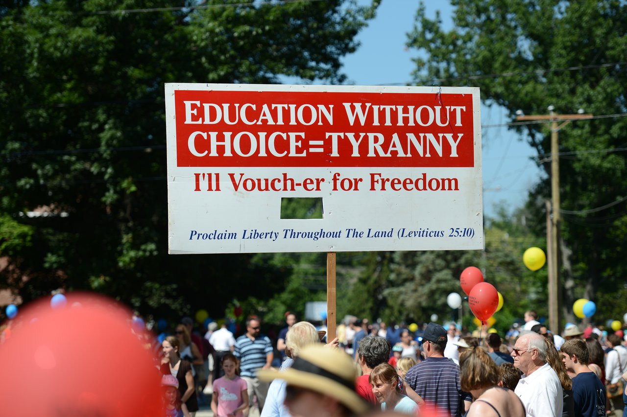 A large sign about education choice is held up during a crowded Fourth of July parade with balloons and spectators.