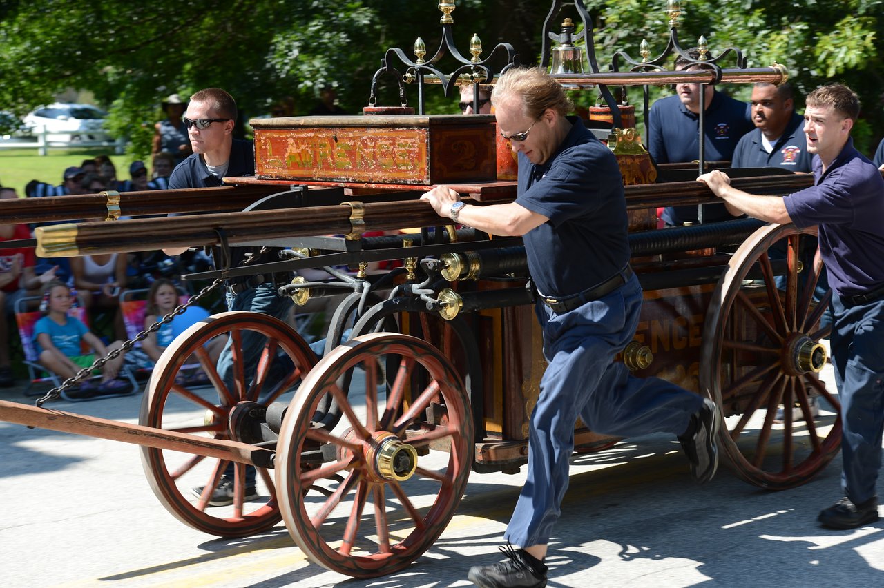 Men in matching uniforms push an antique fire wagon down the street during a parade, with spectators watching.