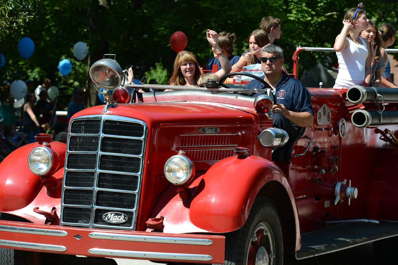 A red vintage fire truck drives in a parade, with people on board waving to the crowd.