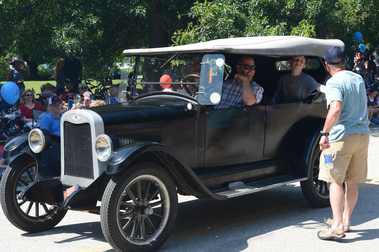 A vintage car drives in a parade while passengers smile and talk to a man standing nearby.
