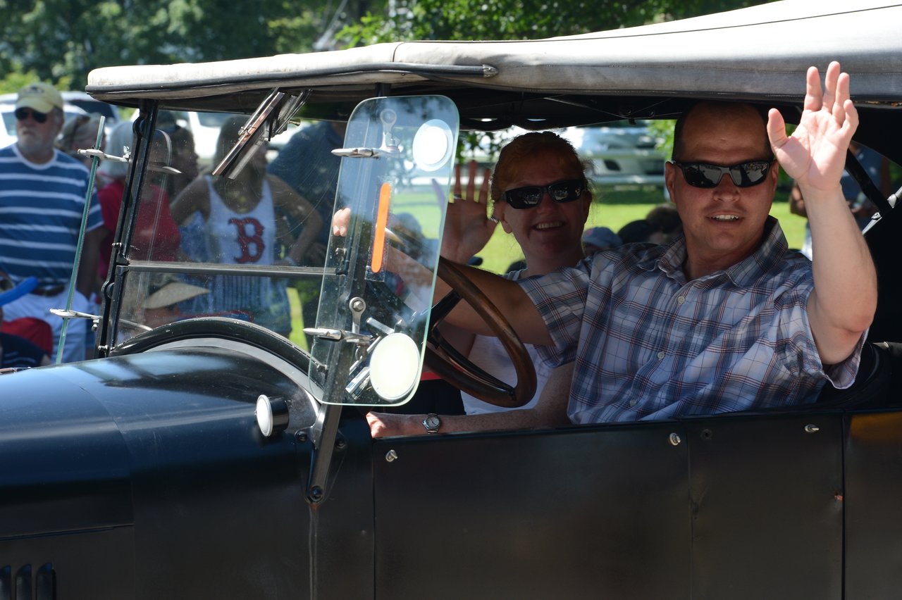 Two people in an old-fashioned car wave to the crowd during a Fourth of July parade.