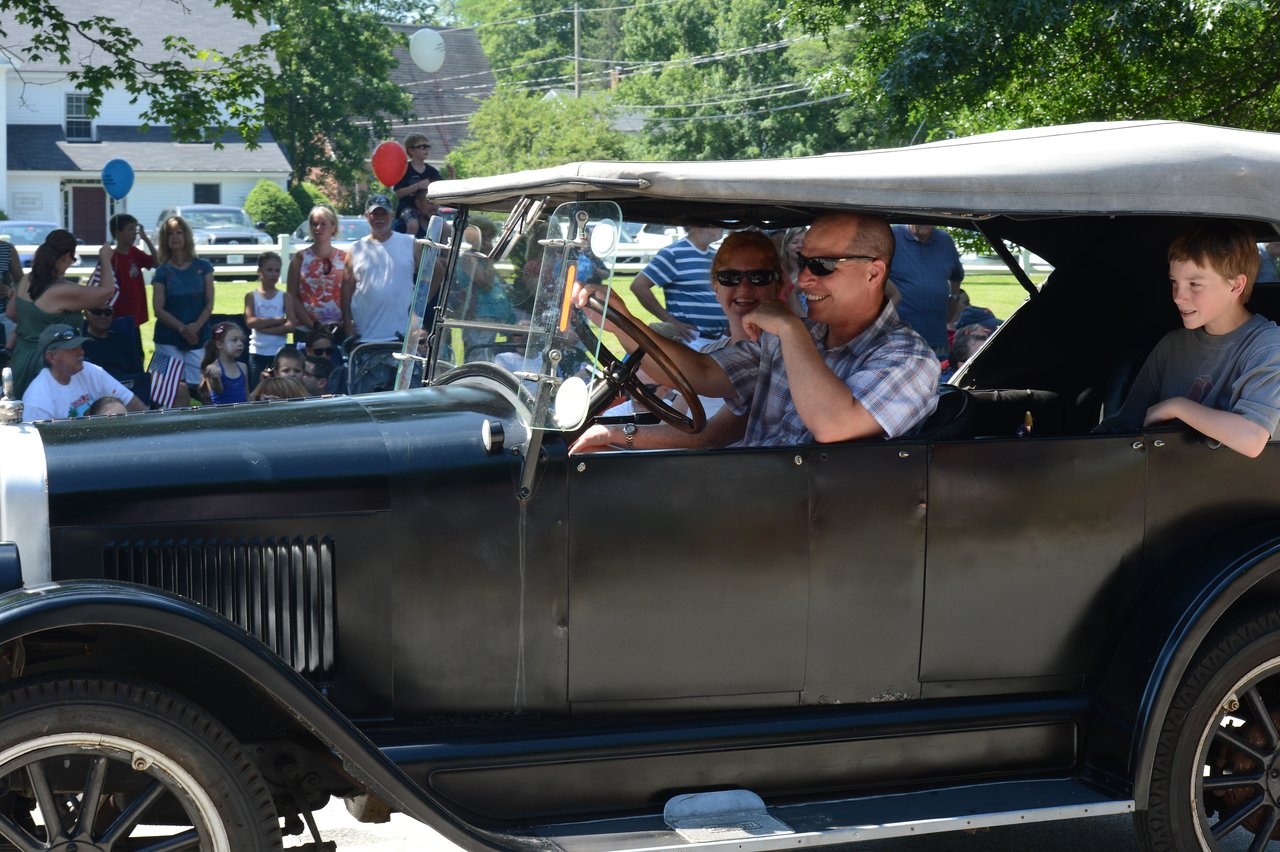 A man drives a vintage car in a parade, smiling and speaking into a microphone, with spectators watching.