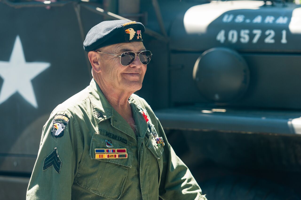 A veteran in a military uniform walks in the 4th of July parade, with an army vehicle behind him.