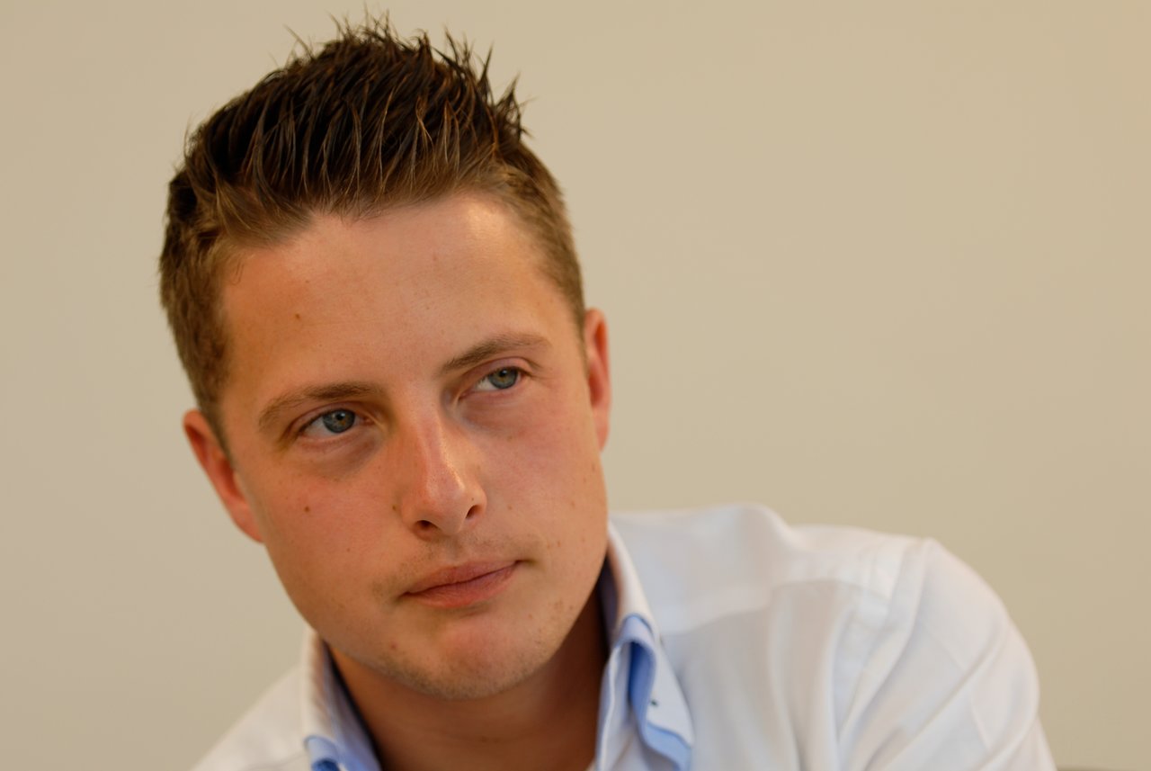 A young man in a white shirt looks off to the side with a neutral expression.