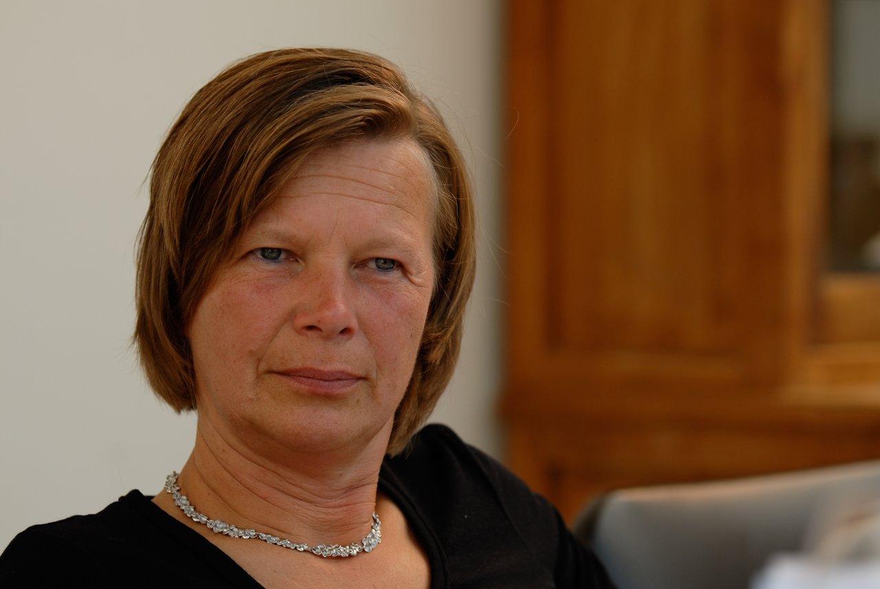 A woman with short brown hair and a necklace sits indoors, looking directly at the camera with a neutral expression.