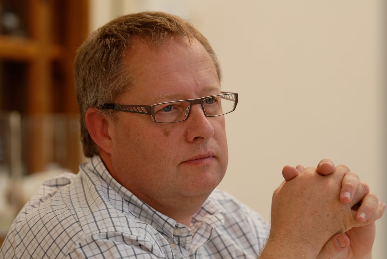 A man wearing glasses and a checkered shirt sits with his hands clasped, looking thoughtful at a gathering.