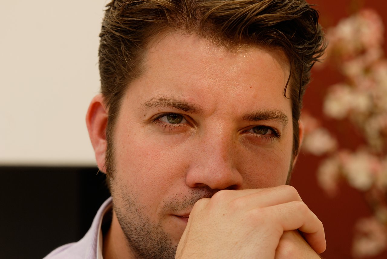A close-up of a man with short, wavy brown hair and light stubble, resting his chin on his hand, with a blurred background featuring flowers and a red wall.