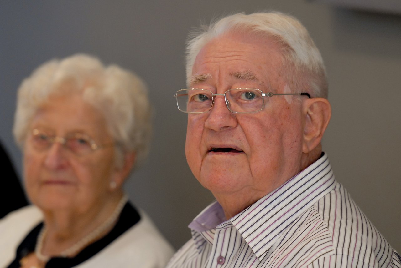An elderly man with white hair, glasses, and a striped shirt looks slightly off-camera, while an elderly woman with white hair, glasses, and a pearl necklace sits blurred in the background.