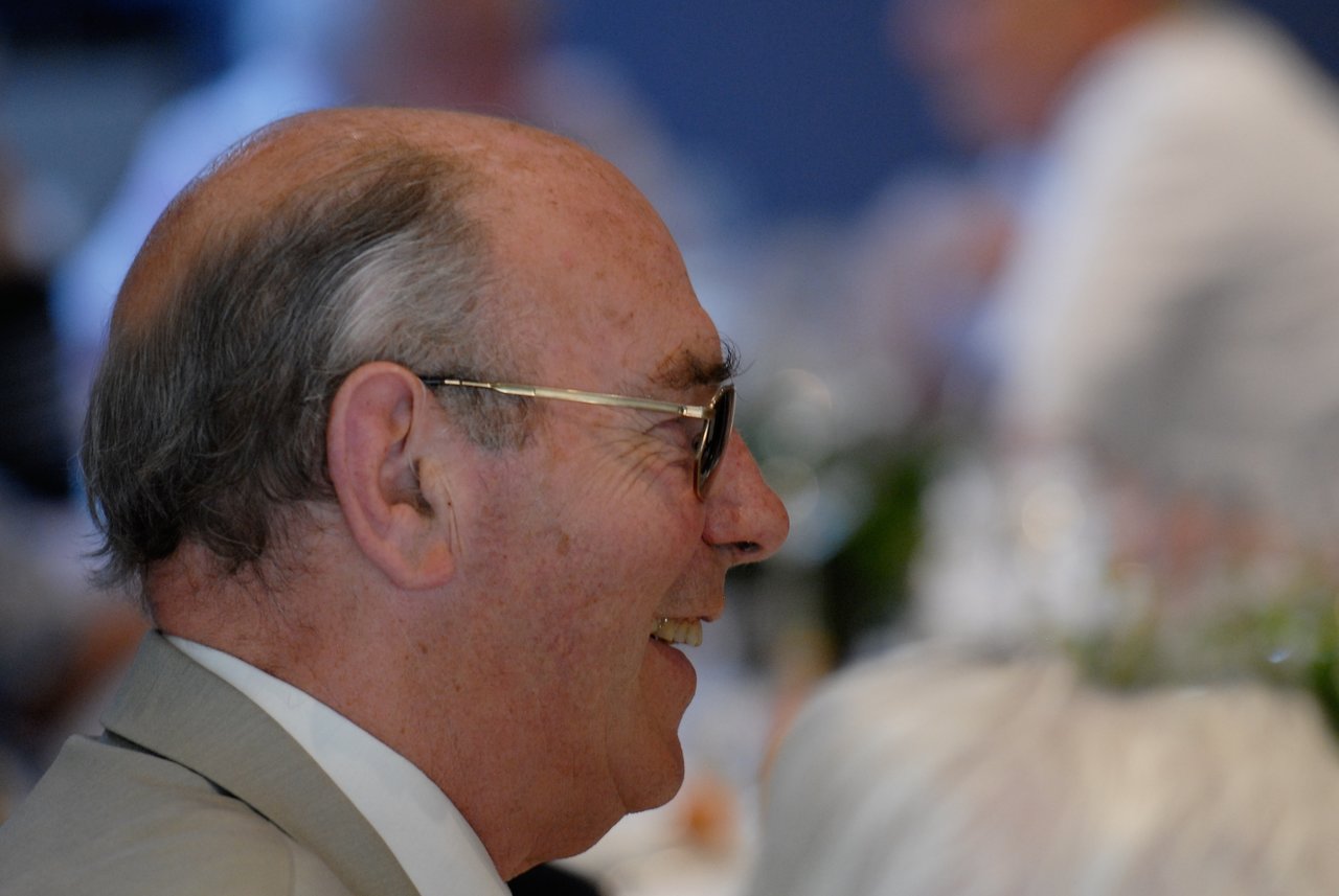 An older man in a suit and glasses smiles while engaged in conversation at a party.