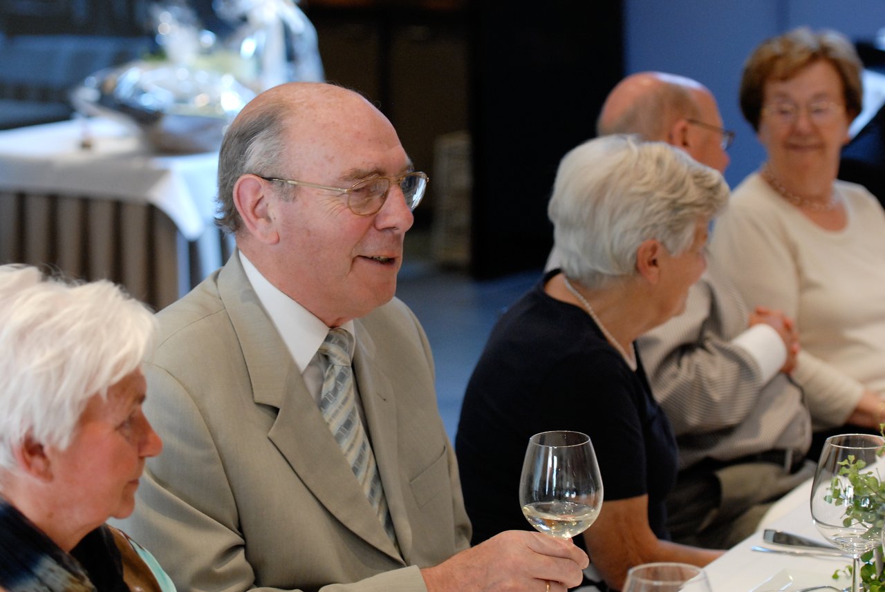 An elderly man in a suit holds a wine glass and smiles while talking with others at a celebration dinner.