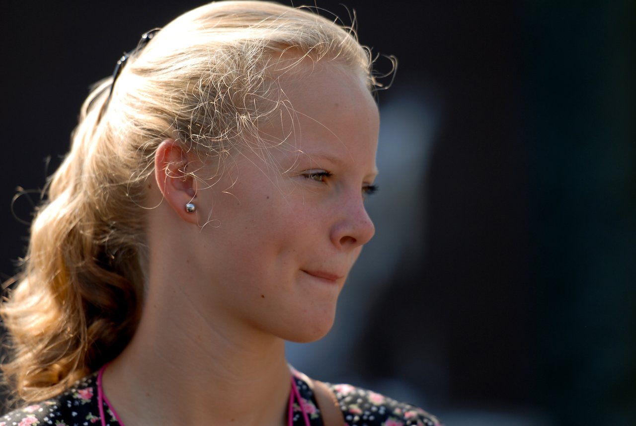 A young woman with blonde hair looks to the side with a neutral expression at an outdoor gathering.