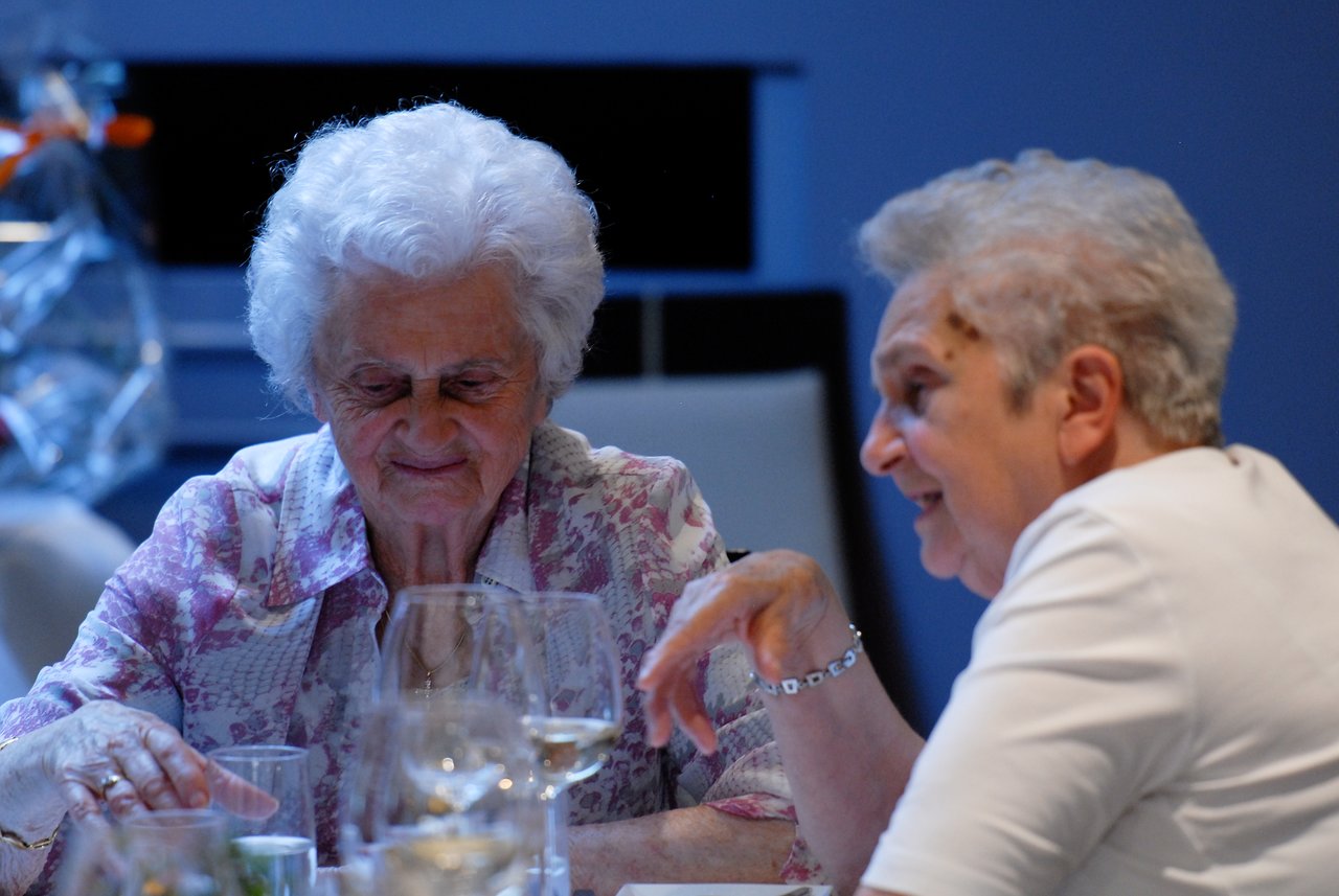 Two elderly women sit at a table, one speaking while the other listens with a smile.
