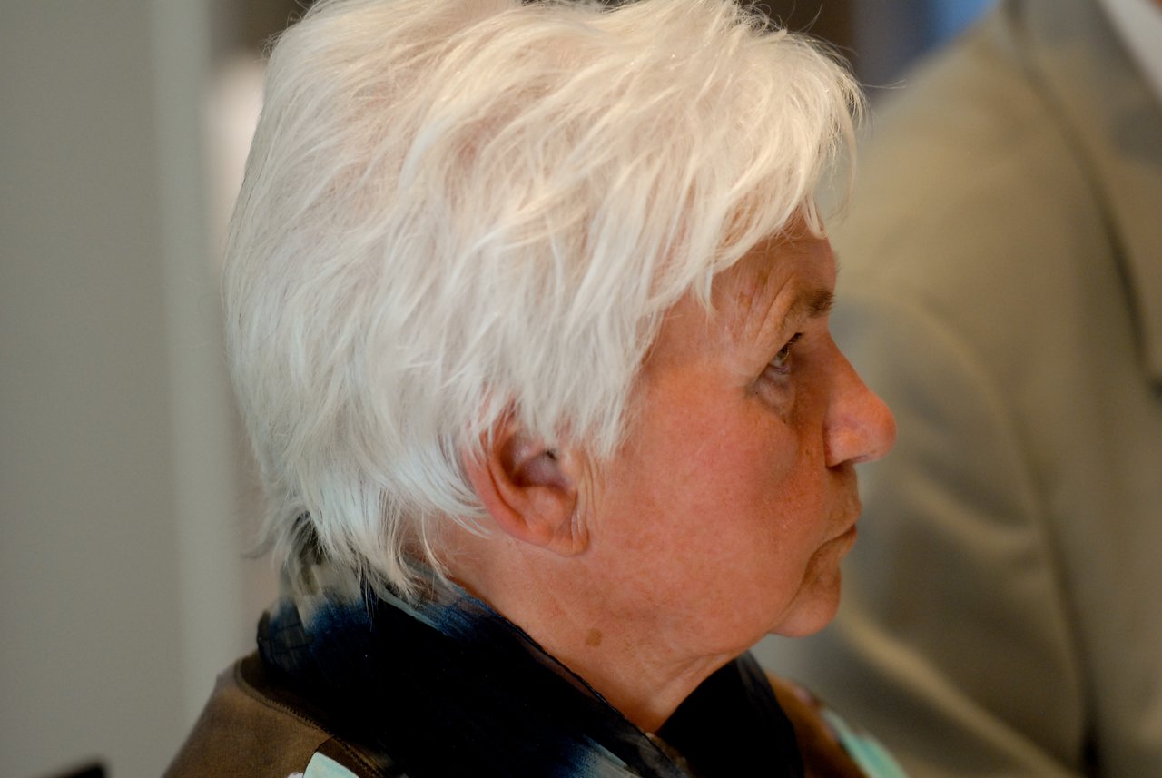 An older woman with short white hair looks to the side at a gathering.