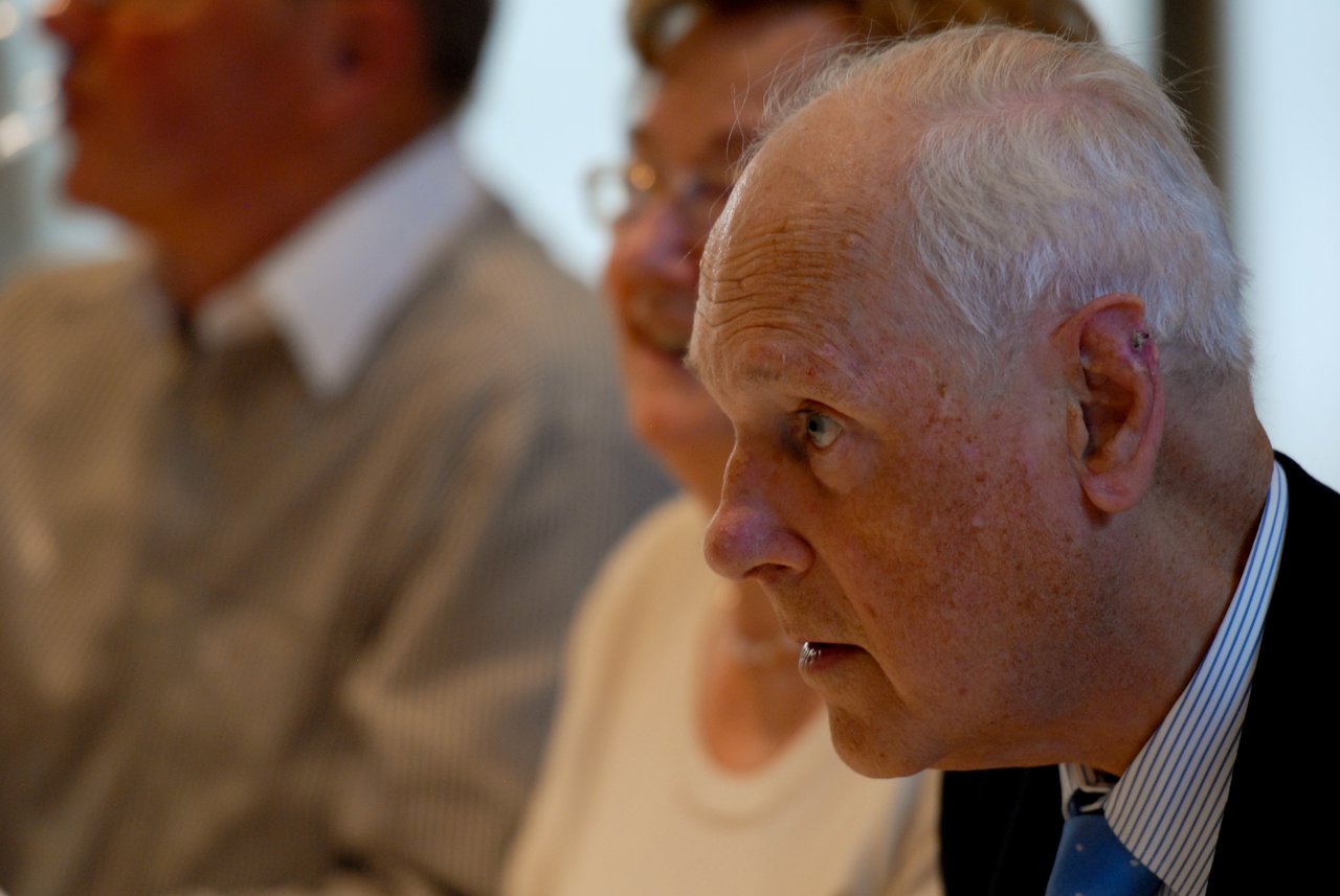 An elderly man in a suit listens attentively at a gathering, with two other people seated behind him.