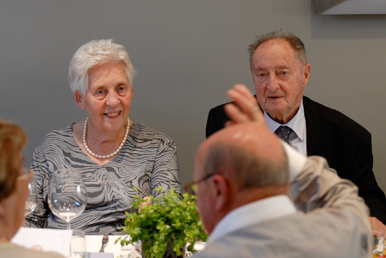 An elderly couple sits at a table, smiling and engaged in conversation during a celebration.