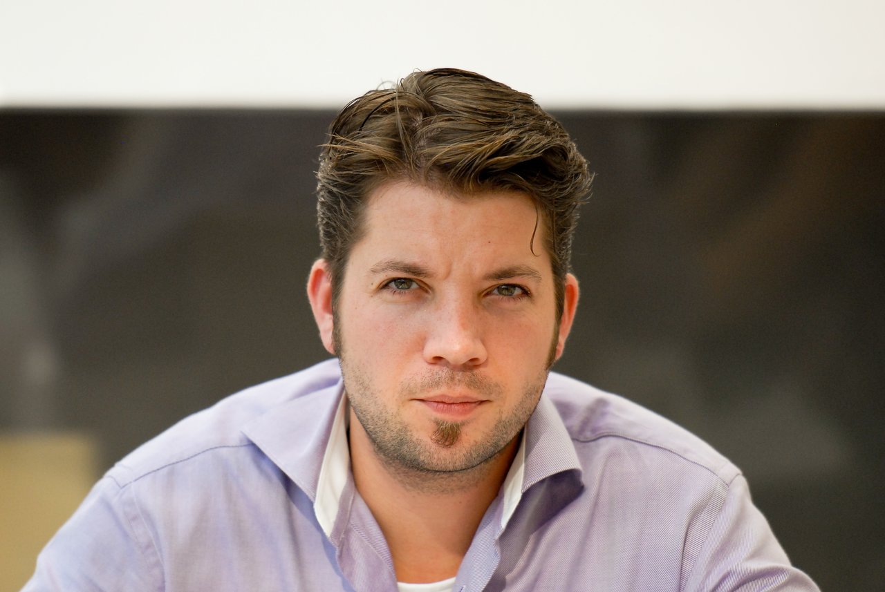 A man in a light purple shirt looks directly at the camera with a neutral expression.