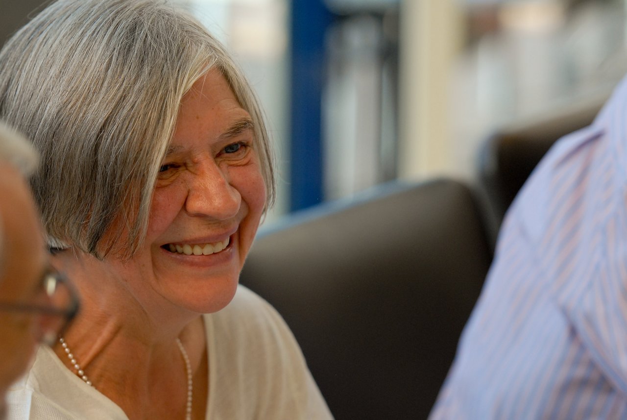 An older woman with gray hair smiles while talking with others at a gathering.