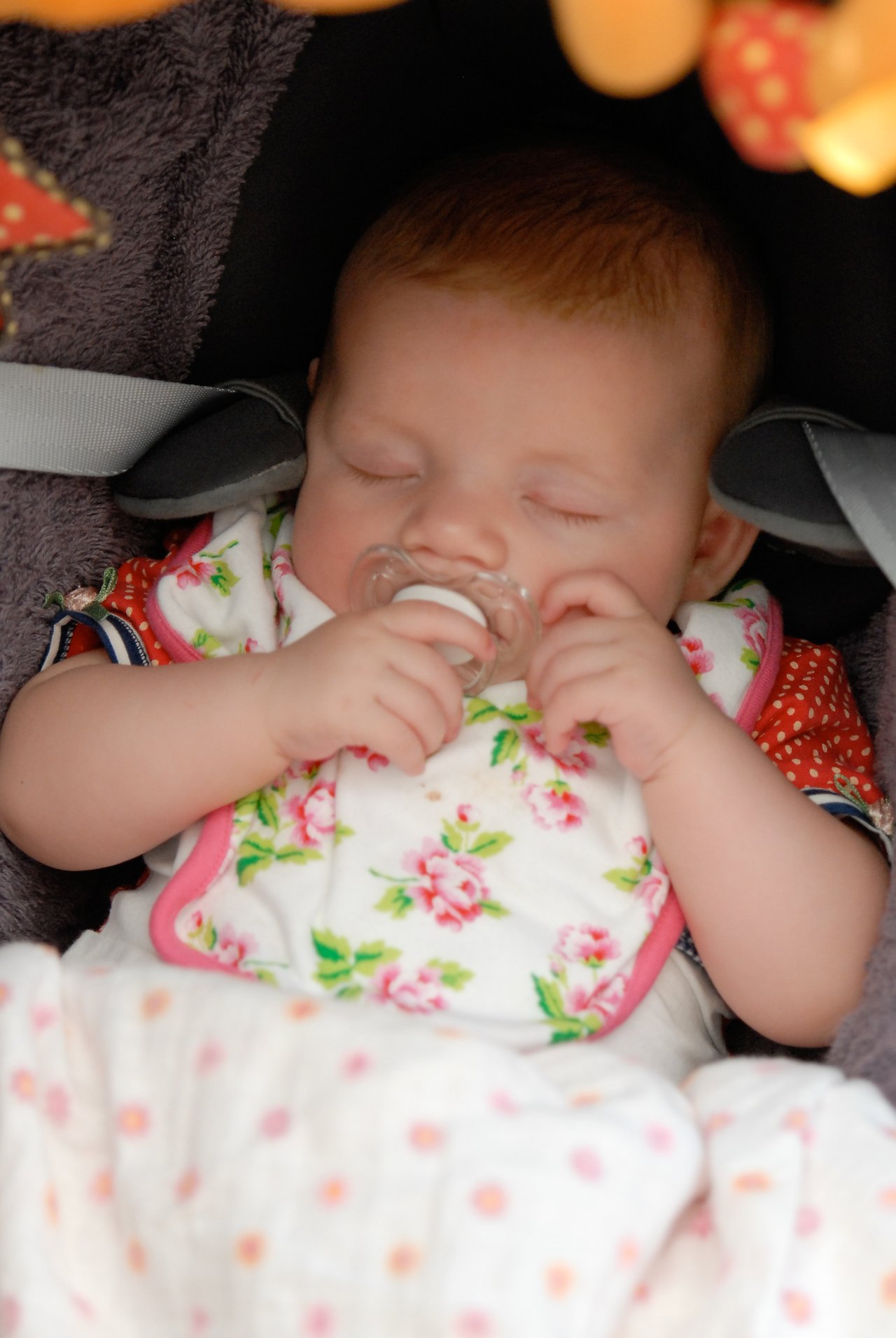 A baby sleeps in a car seat, holding a pacifier, wearing a floral bib and a red outfit.