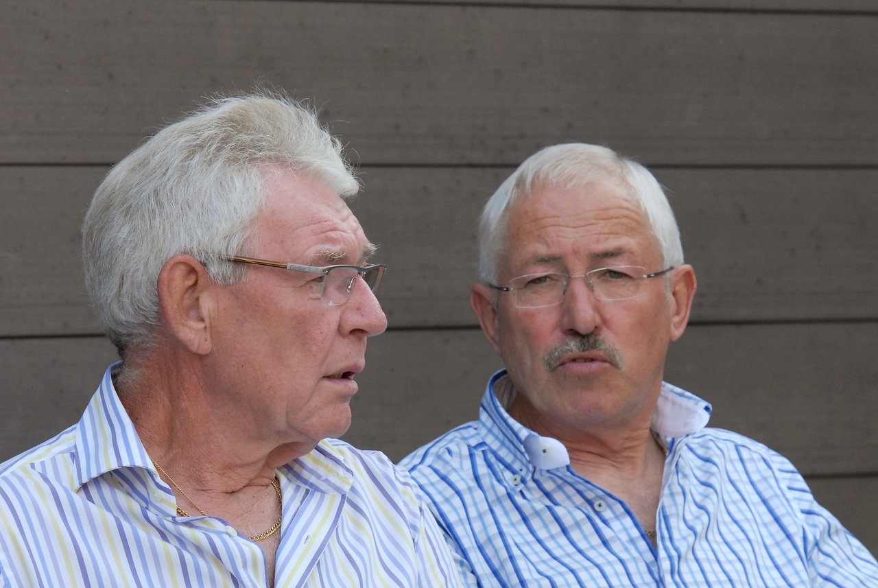 Two older men in striped shirts sit and talk, one speaking while the other listens attentively.