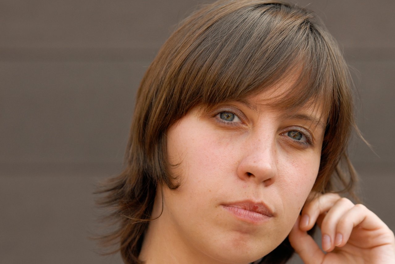 A woman with short brown hair looks at the camera while resting her hand near her ear.