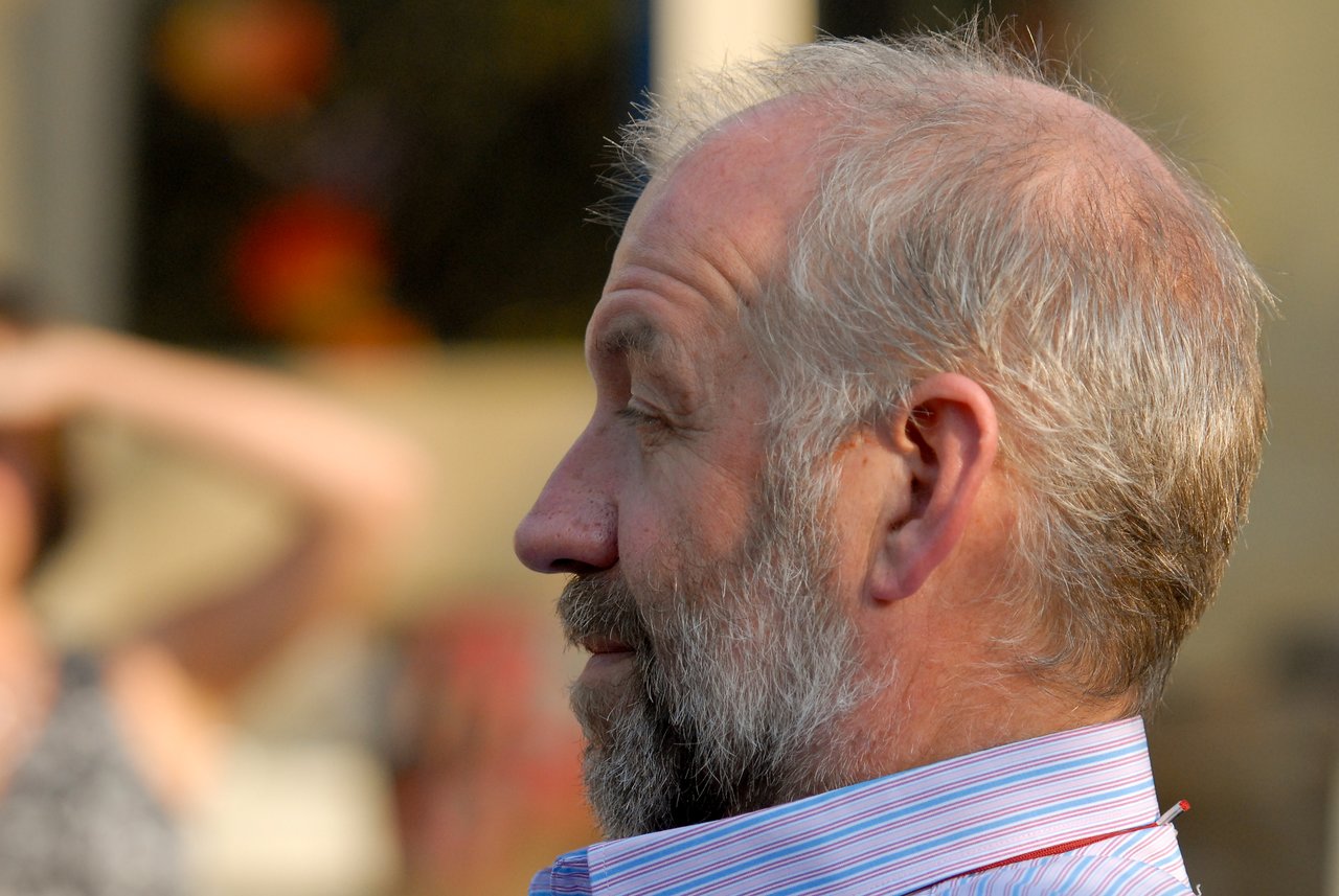 A bearded man in a striped shirt looks to the side at an outdoor gathering.