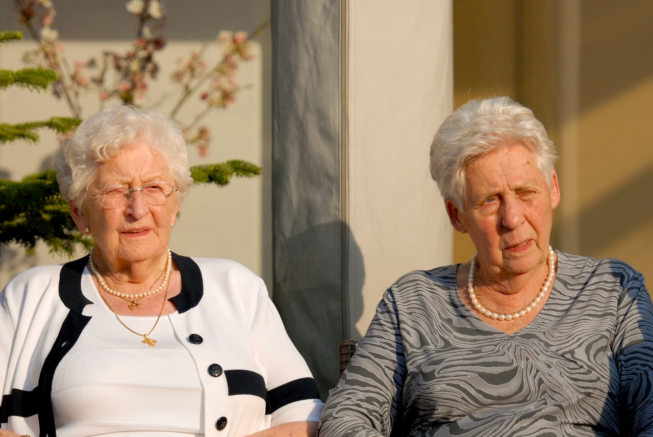Two elderly women sit outdoors in the sunlight, dressed in elegant outfits with pearl necklaces, attending a celebration.