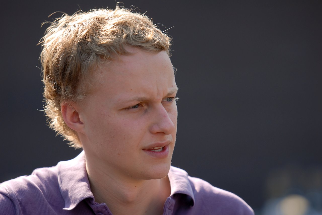 A young man in a purple shirt looks to the side with a focused expression at an outdoor gathering.