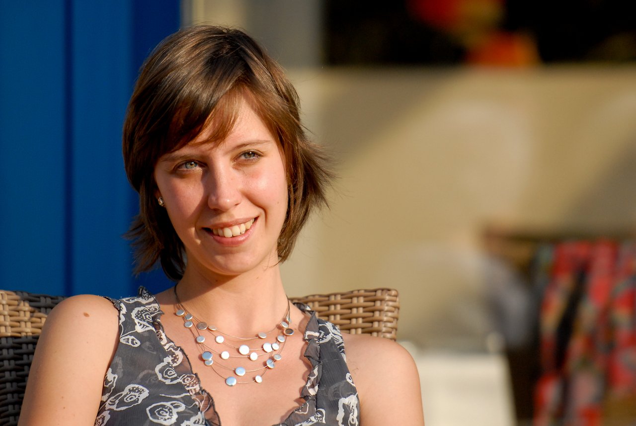 A woman sits on a wicker chair, smiling and looking slightly to the side at a party.