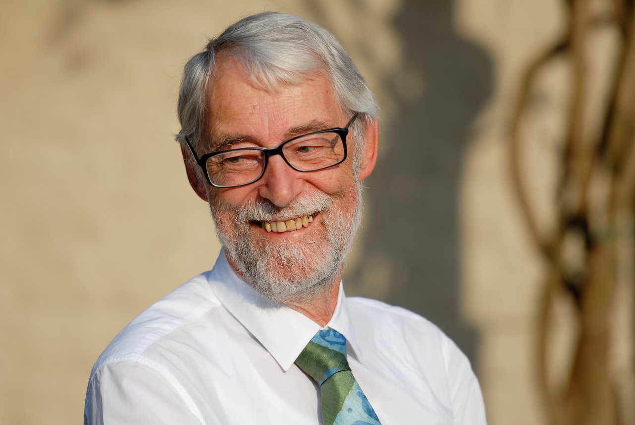 An older man with glasses and a beard smiles while wearing a white shirt and a green tie.
