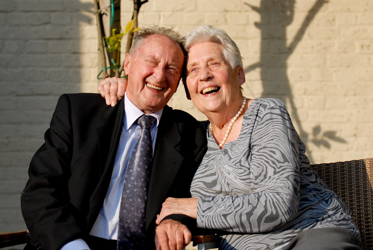 An elderly couple sits closely together, smiling and laughing at a celebration.