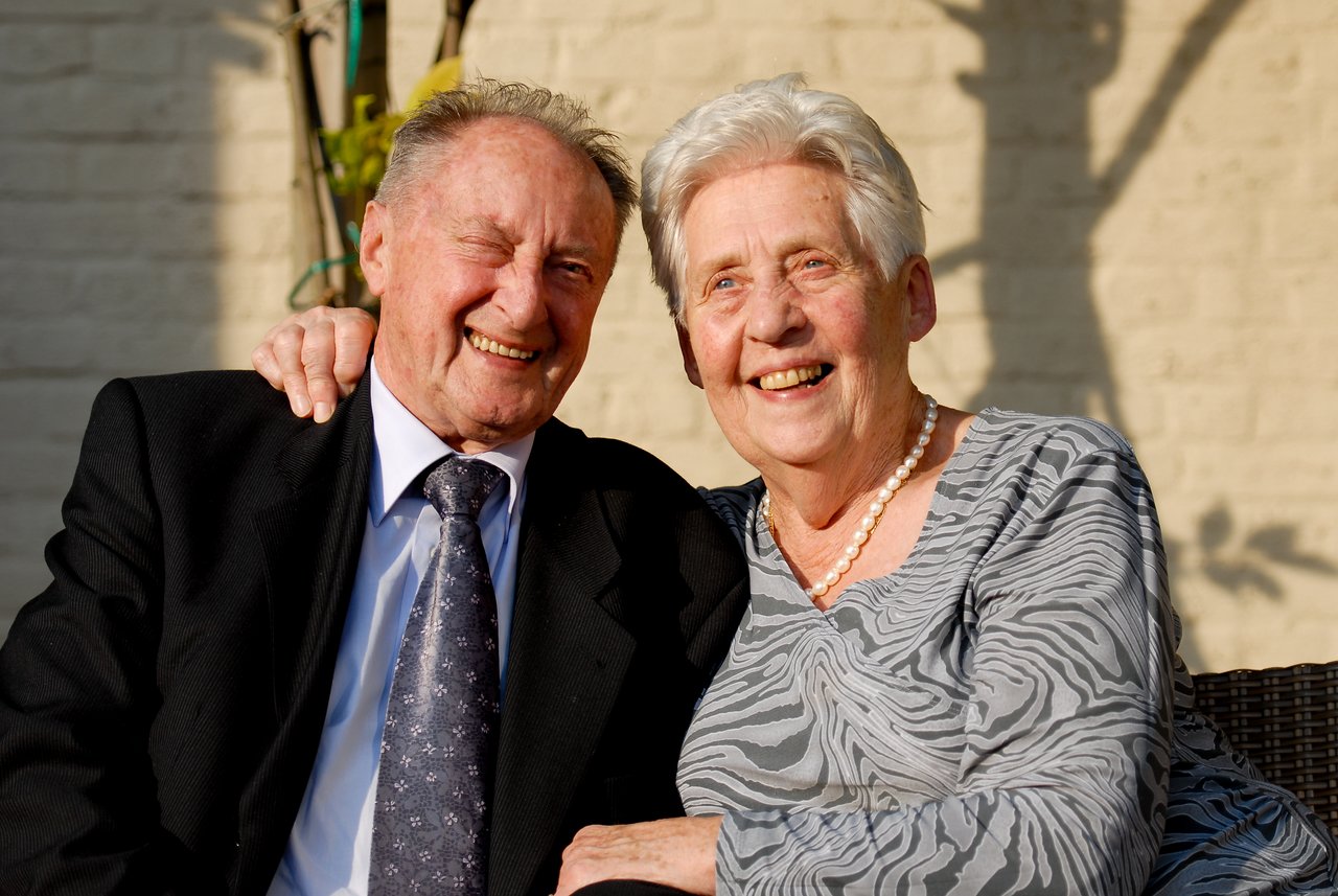 An elderly couple sits closely together, smiling and dressed formally, celebrating their 60th anniversary at a party.