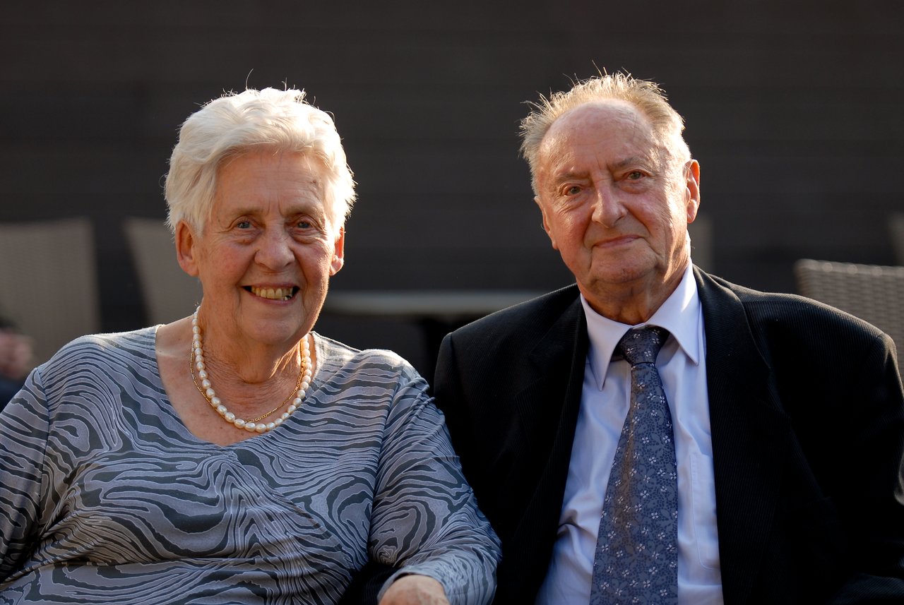 An elderly couple sits together, smiling at the camera during a celebration.