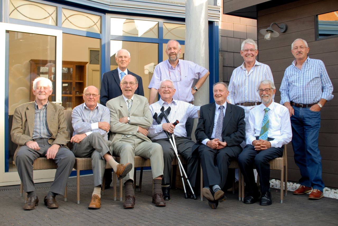 A group of older men pose together outside, smiling and dressed in formal and casual attire.