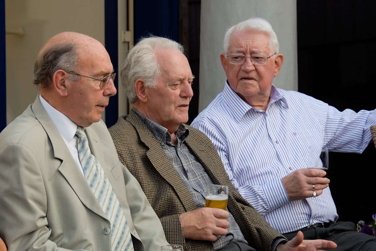 Three older men sit together, talking and holding drinks at a party.