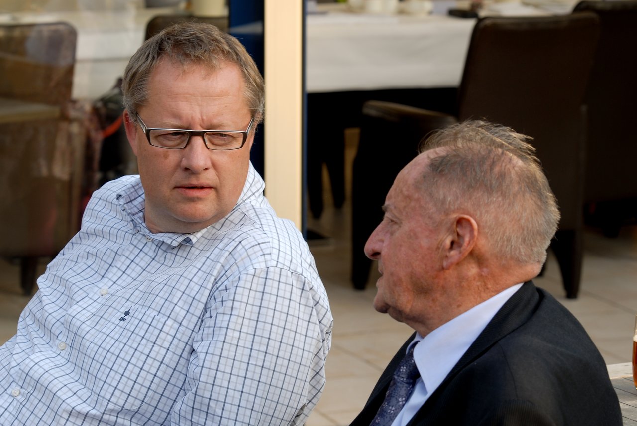 Two men sit and talk at a party, one wearing glasses and a checkered shirt, the other in a suit.