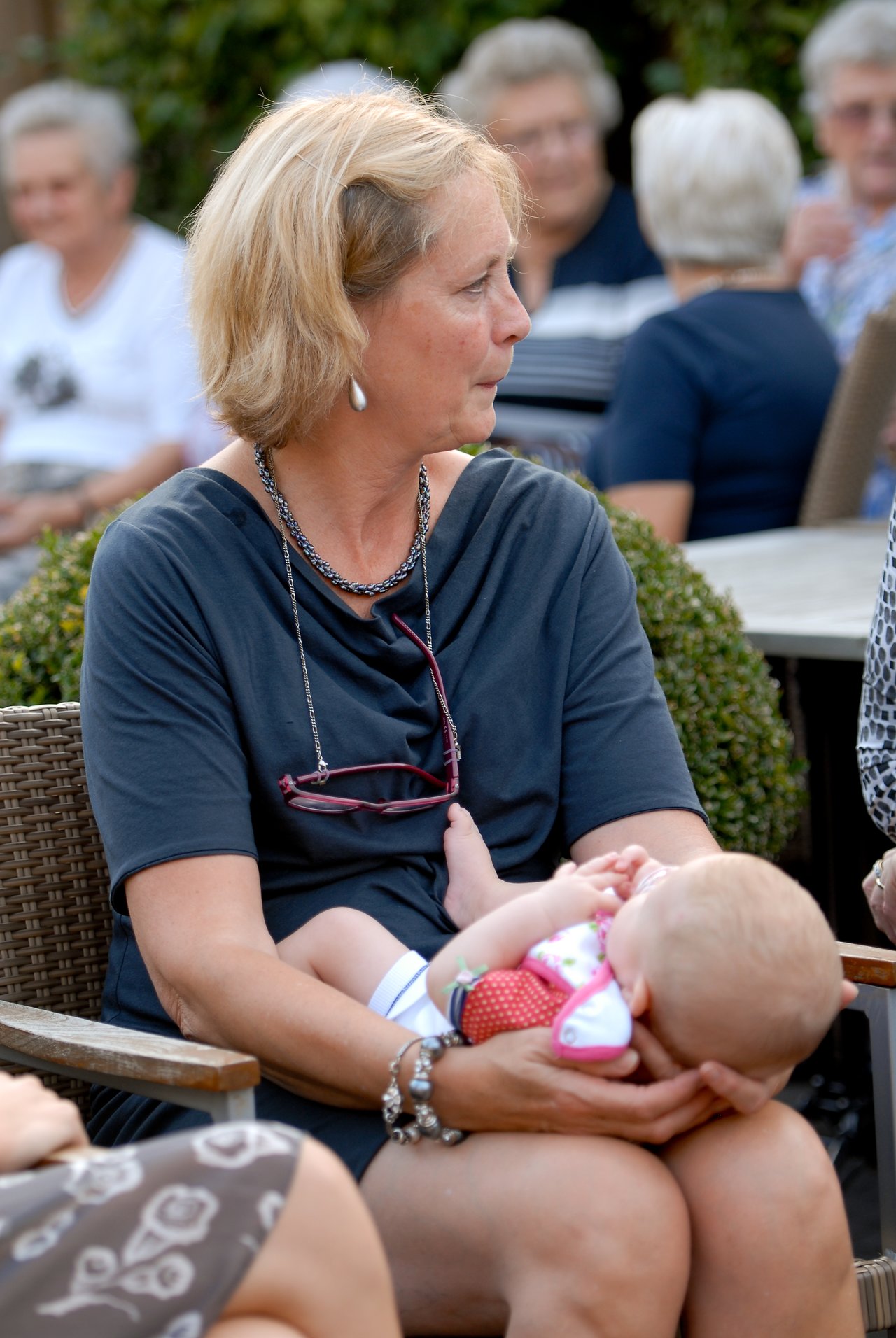 A woman sits holding a baby in her arms while looking to the side at an outdoor gathering.