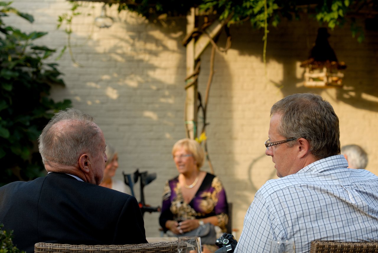Two men sit at a table, engaged in conversation, while a woman in the background smiles and talks.