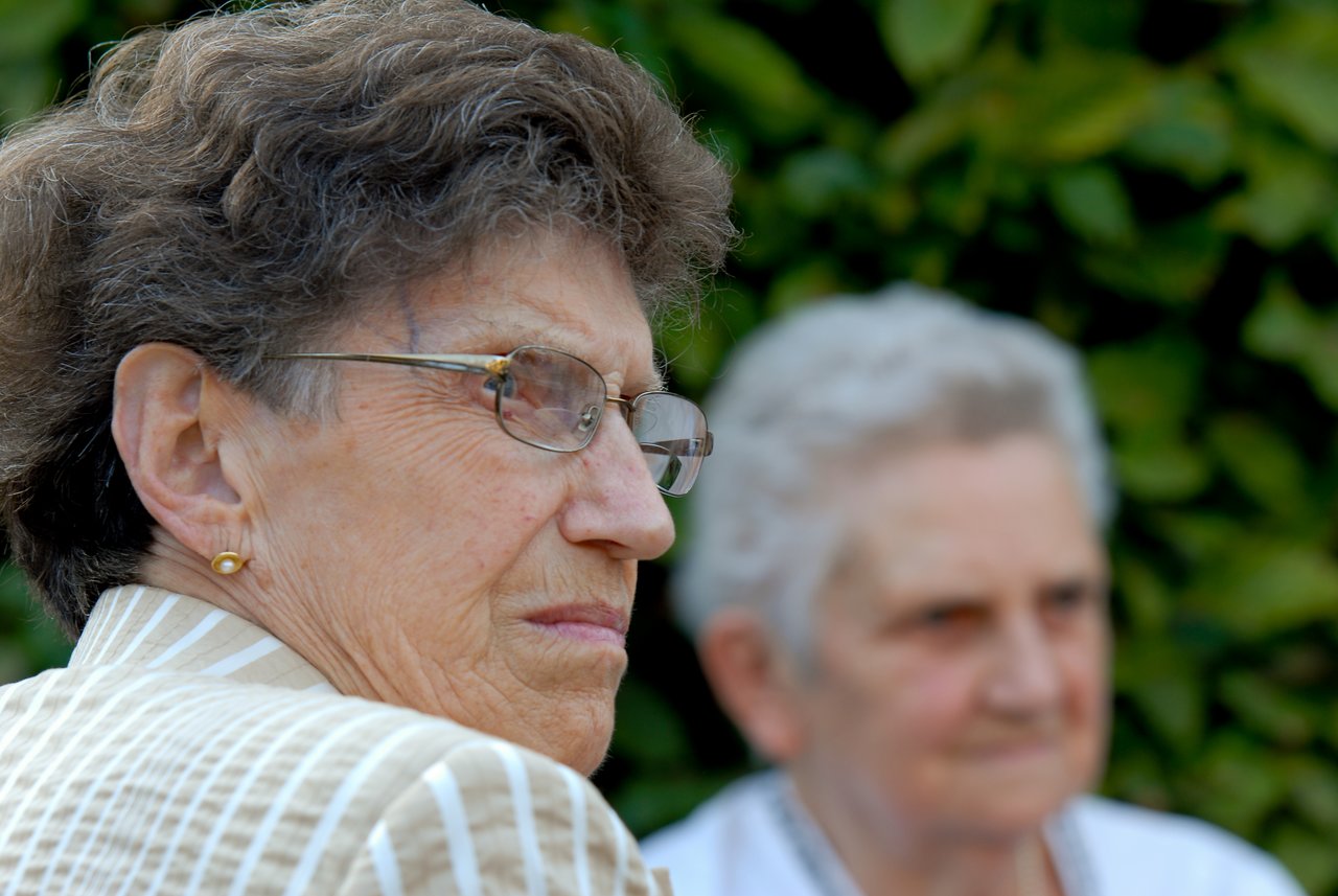 Two elderly women sitting outdoors, one in focus wearing glasses and looking to the side, the other slightly blurred.