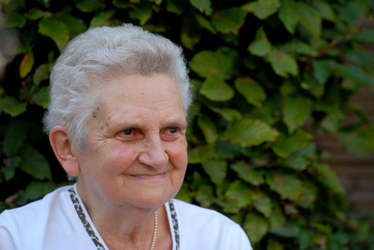 An elderly woman with short gray hair smiles while looking off to the side, wearing a white shirt and necklace.