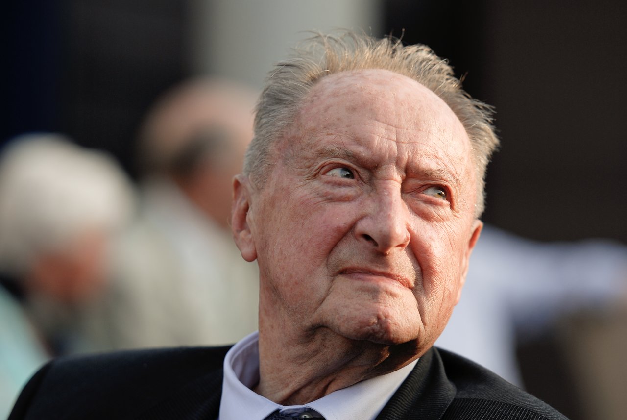 An elderly man in a suit looks off to the side at a gathering, with blurred people in the background.