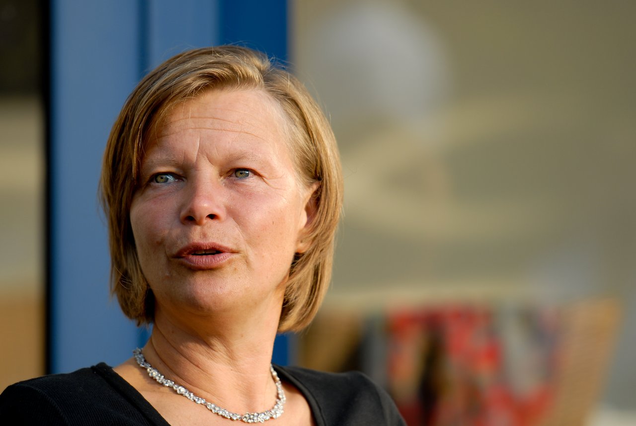 A woman in a black top and necklace is speaking at a party, looking slightly off-camera.
