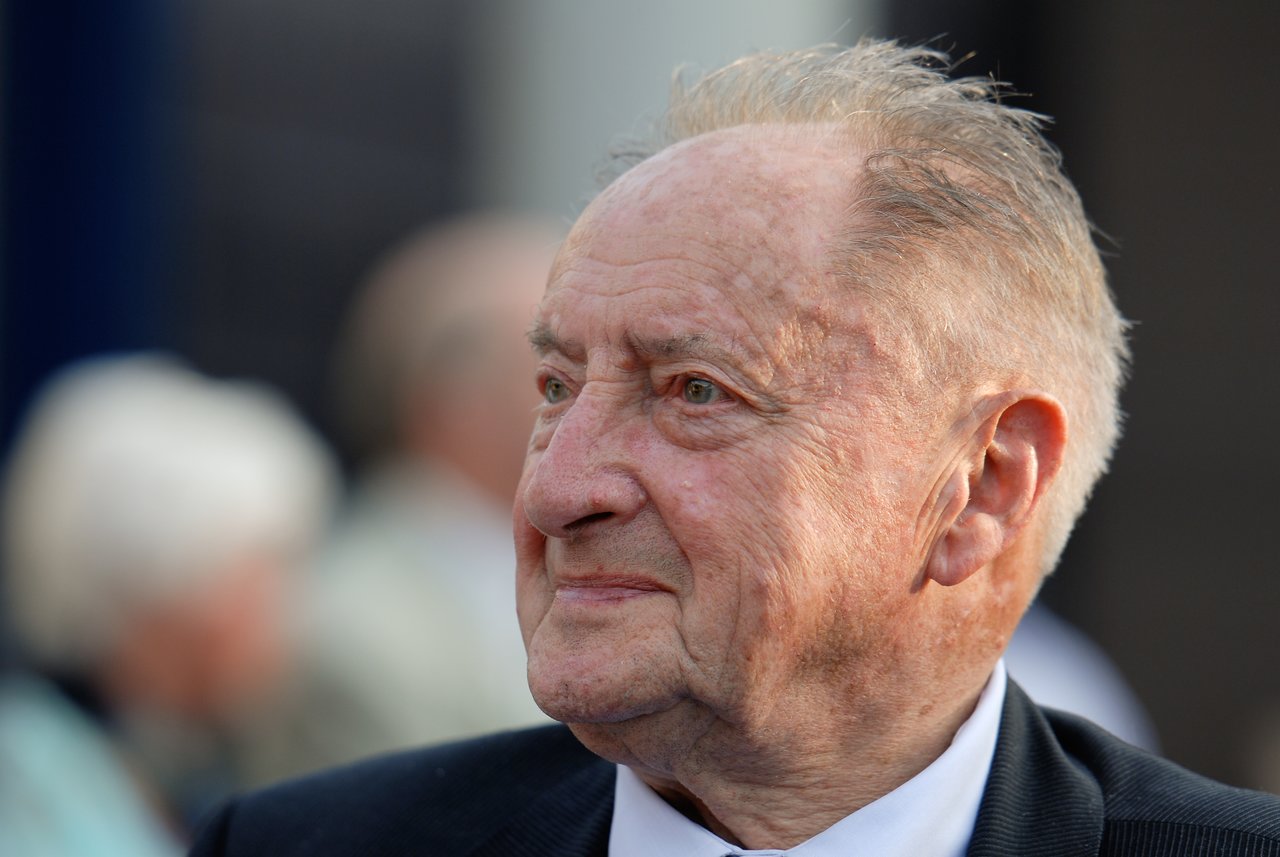 An elderly man in a suit smiles while looking off to the side at a party.