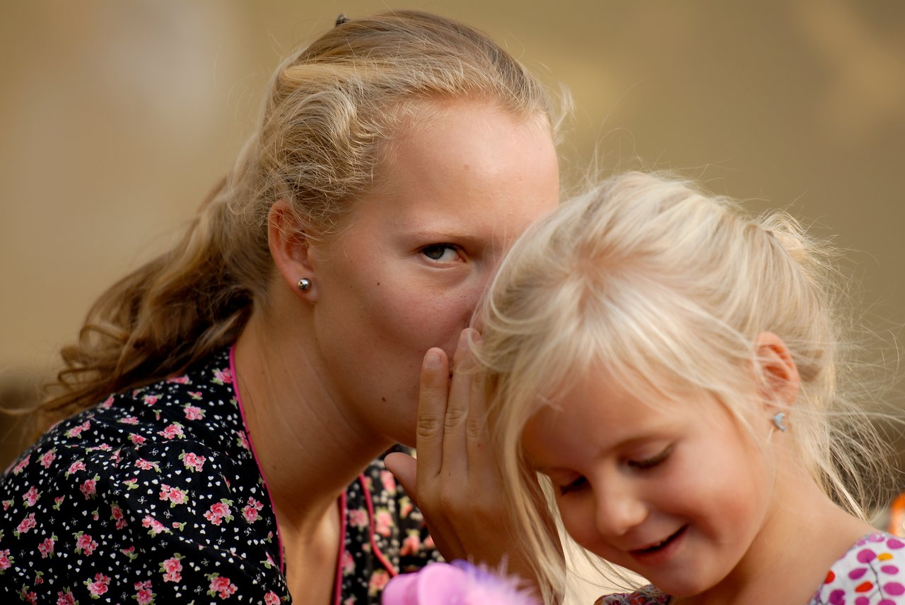 A woman whispers into a young girl's ear while the girl smiles and looks down.