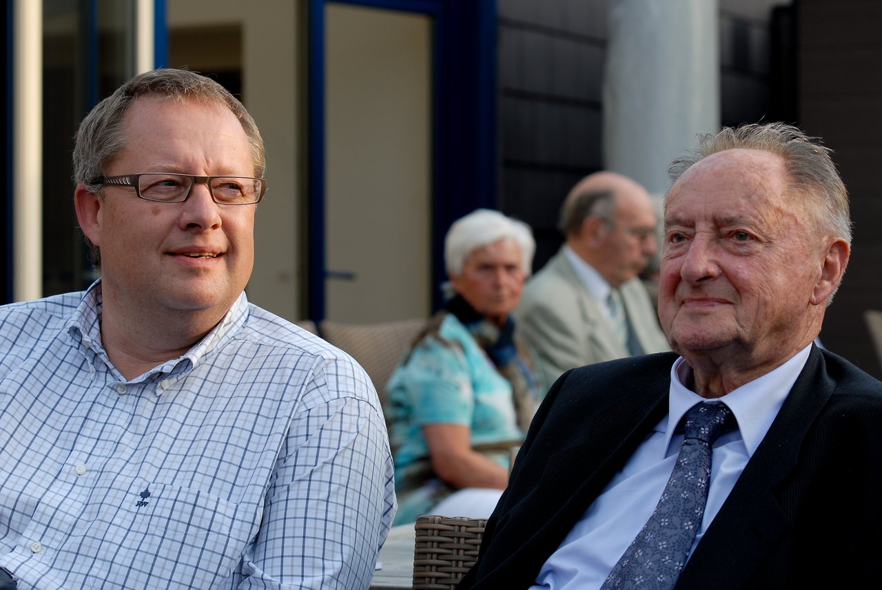 Two men sit together at an outdoor gathering, smiling and engaged in conversation.