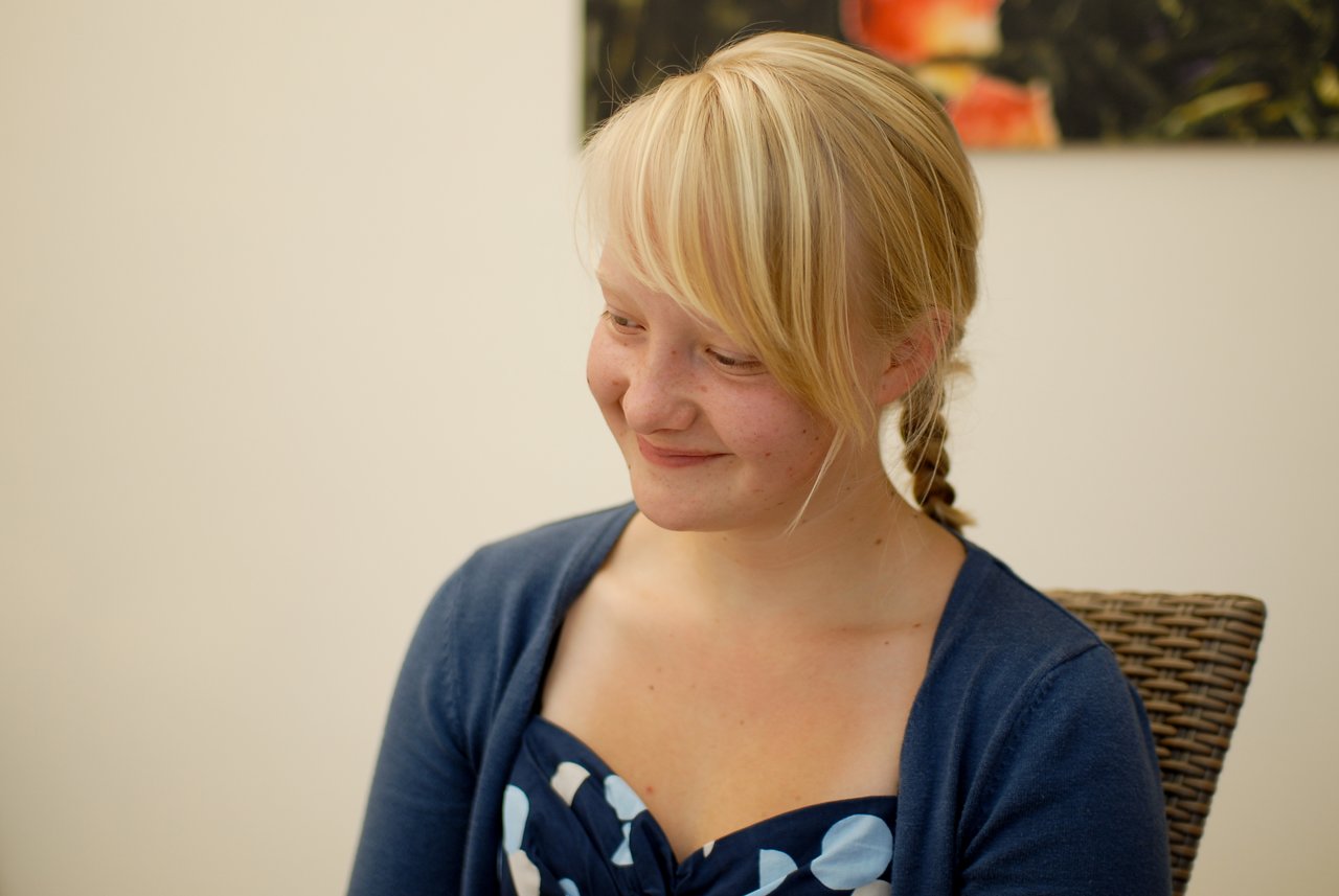A young woman with blonde hair smiles while sitting at a party, wearing a blue dress and cardigan.