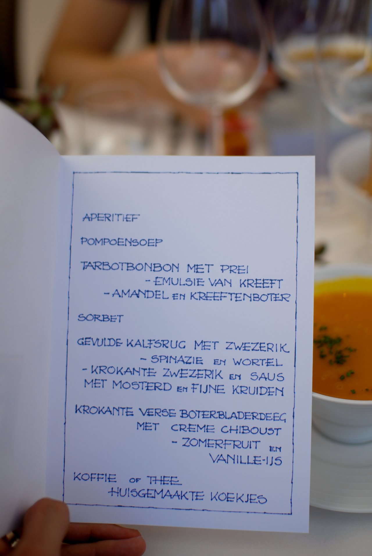 A person holds a menu with Dutch text while a bowl of pumpkin soup sits on the table.