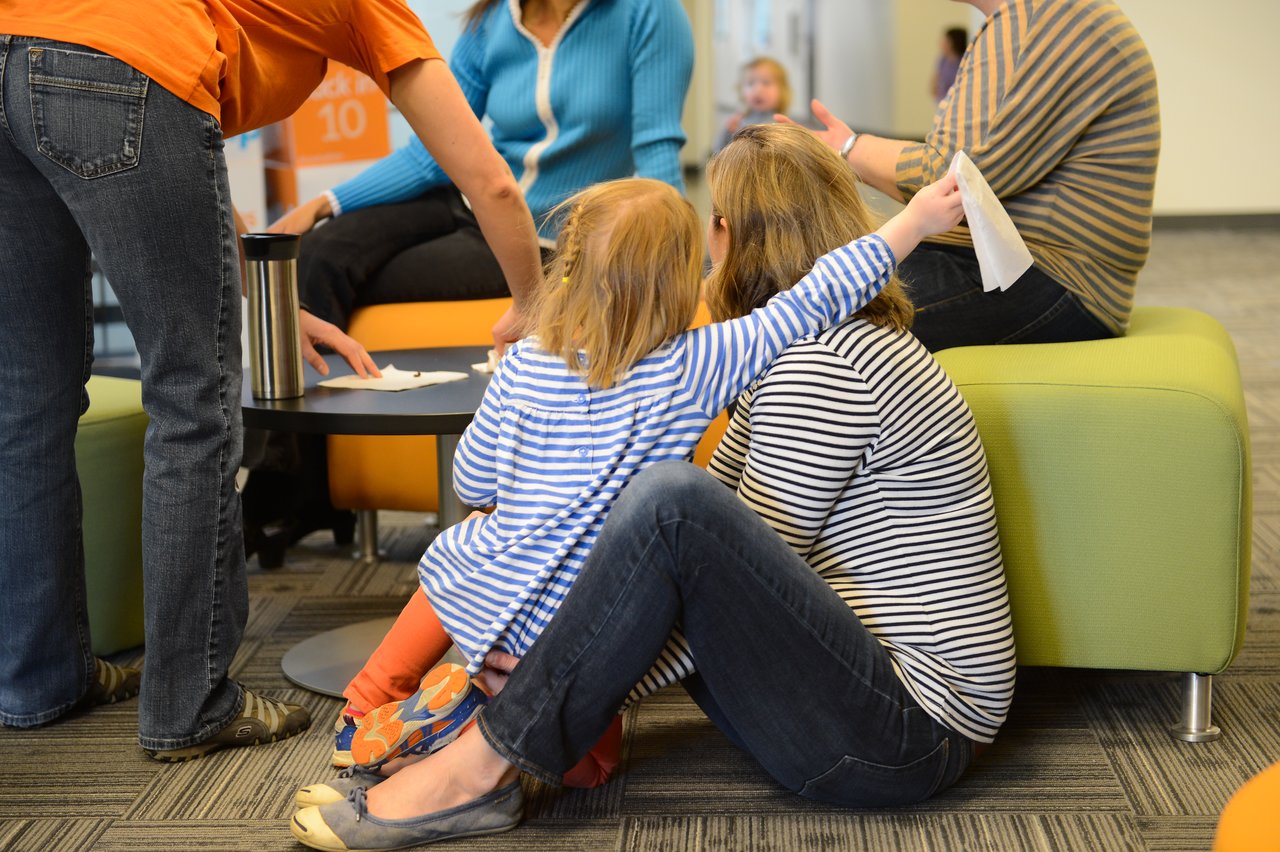A child in a striped shirt sits on a woman's lap, reaching out with a tissue in hand.