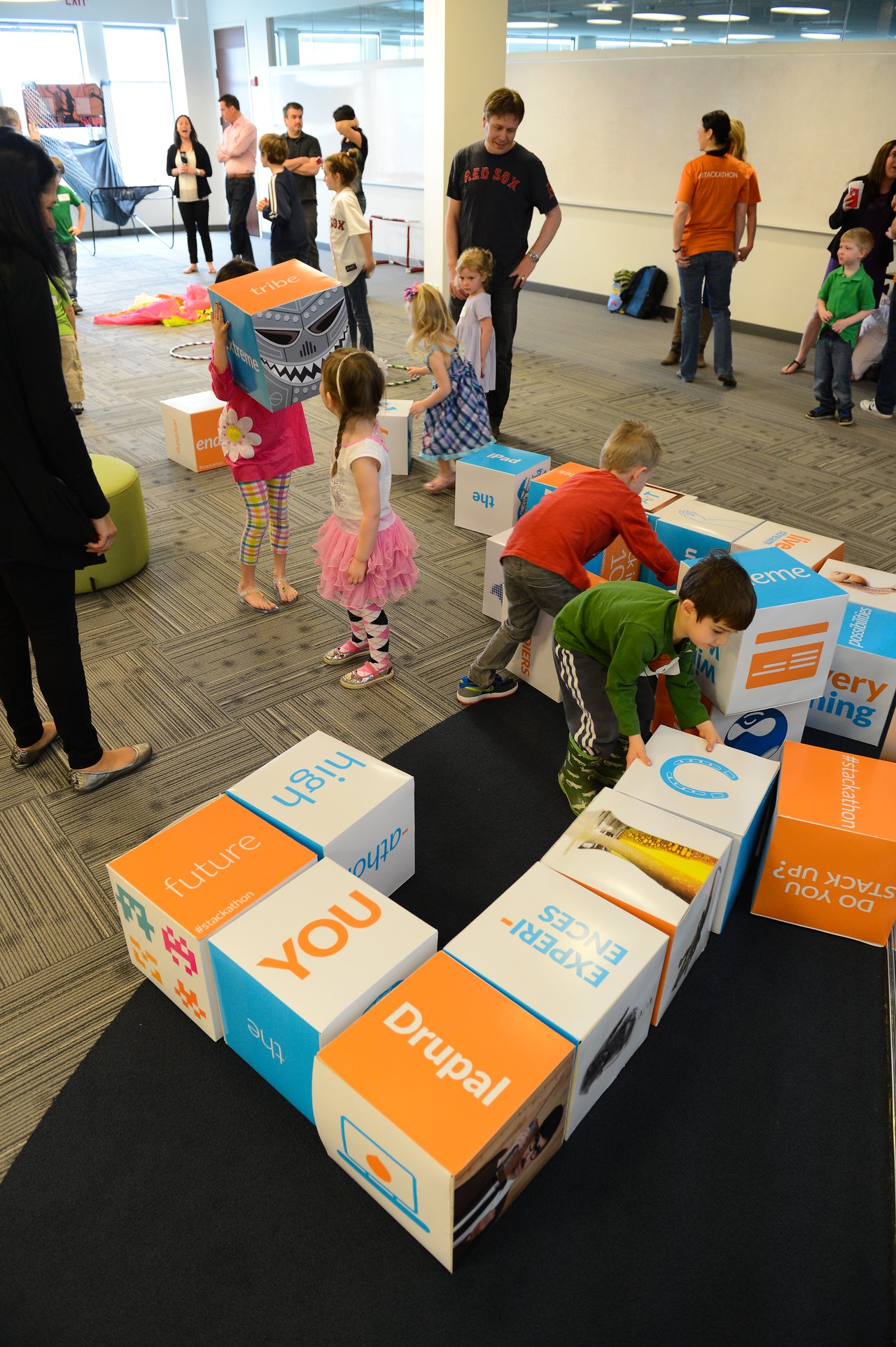 Children play with large branded blocks in an office space while adults observe and interact during a company event.